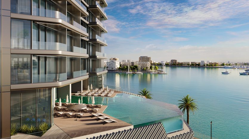 Luxueux immeuble de grande hauteur en bord de mer à Dubaï avec balcons donnant sur une marina remplie de bateaux, doté d&#039;une piscine à débordement et de transats.