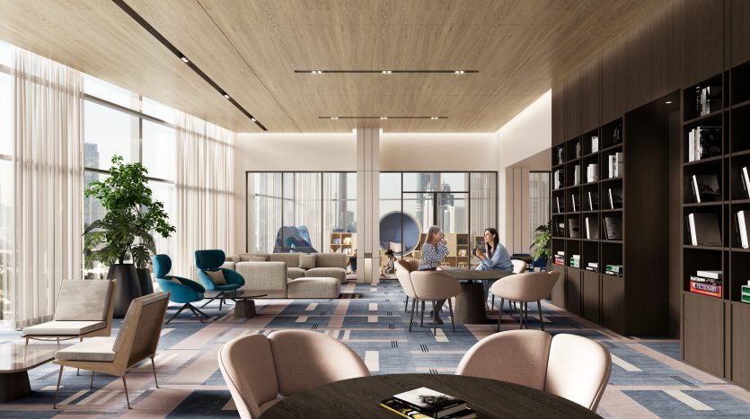 Un salon de bureau moderne à Dubaï avec de hautes fenêtres, de la lumière naturelle et un mobilier élégant comprenant des canapés et des chaises longues. Trois personnes conversent près d’une étagère, entourées de plantes d’intérieur.