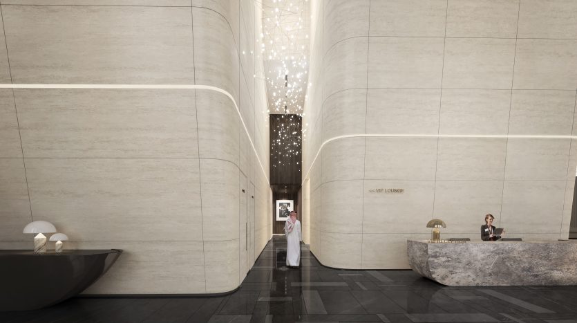 Un hall d&#039;hôtel moderne à Dubaï avec de hauts plafonds incurvés et d&#039;élégants murs en marbre. Un lustre central descend et un homme en tenue traditionnelle passe, accueilli par un réceptionniste assis à un bureau en pierre.