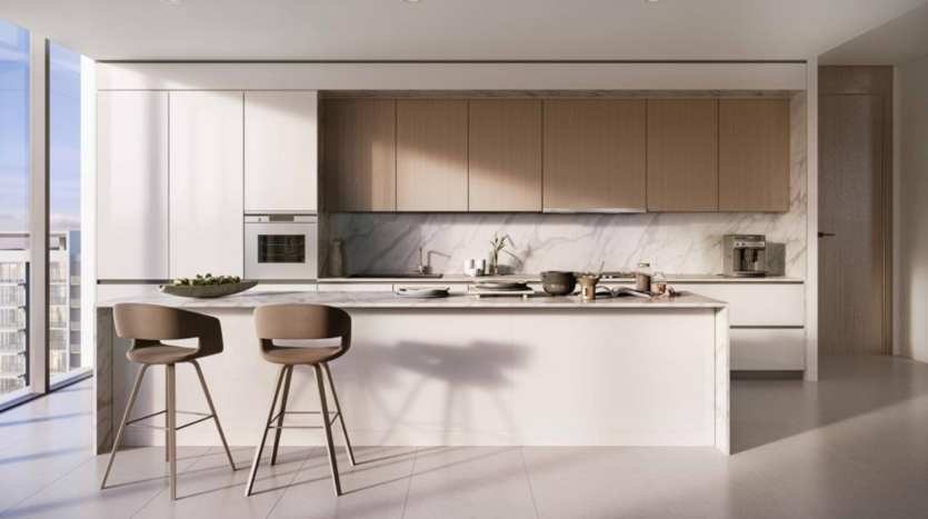 Une cuisine moderne avec des comptoirs en marbre blanc, des armoires en bois, des appareils électroménagers intégrés et un bar de petit-déjeuner avec deux tabourets. La pièce est baignée de lumière naturelle dans un appartement Dubaï.