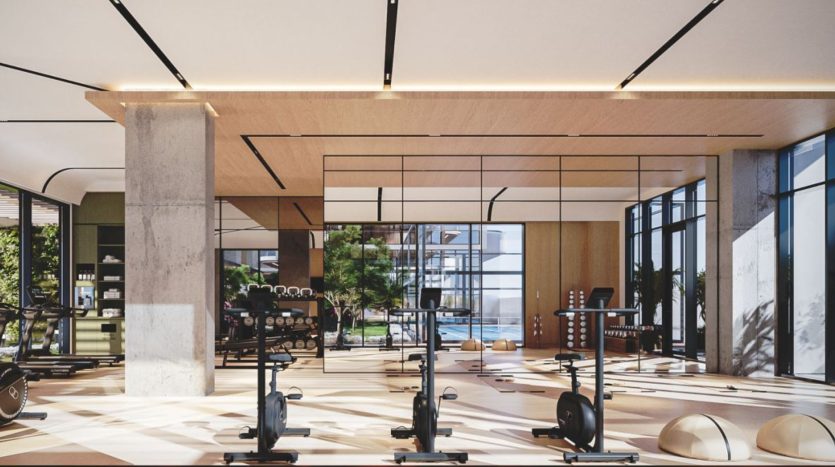 Intérieur de salle de sport moderne comprenant divers équipements d'exercice comme des tapis roulants et des poids, une lumière naturelle abondante et une vue sur la verdure à l'extérieur à travers de grandes fenêtres. Parfait pour ceux qui investissent dans un style de vie actif à Dubaï.