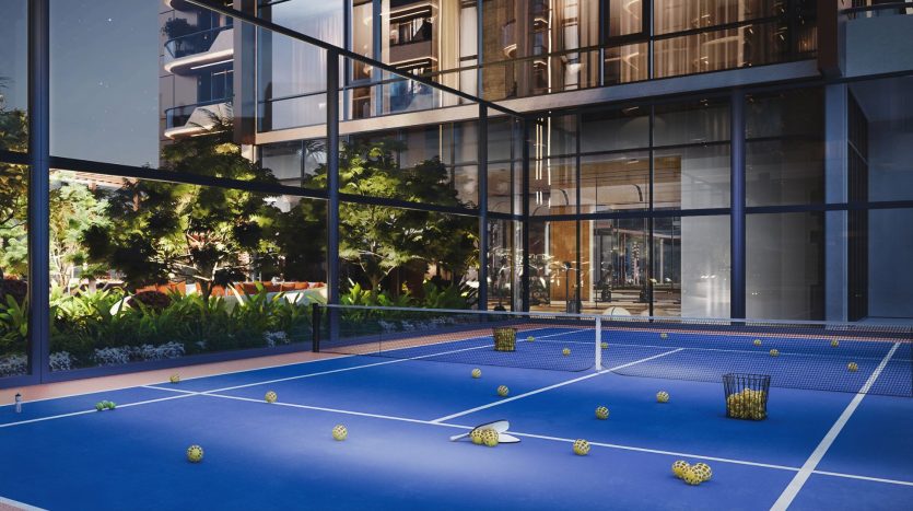 Un court de tennis extérieur avec plusieurs paniers de balles de tennis éparpillés, situé entre des bâtiments modernes aux panneaux de verre ornés de plantes vertes luxuriantes, mettant en valeur un élément privilégié de l'immobilier Dubaï.