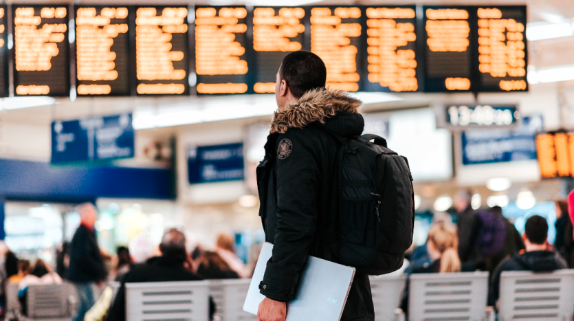 Un homme avec un sac à dos et un ordinateur portable sous le bras se tient face à un grand panneau d&#039;information de vol dans un terminal d&#039;aéroport très fréquenté, planifiant son voyage pour visiter un appartement à Dubaï.