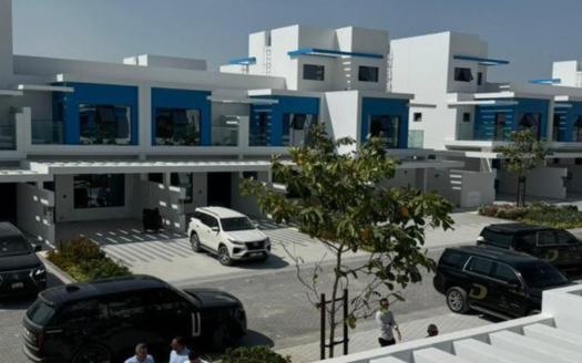 Un quartier résidentiel moderne de Dubaï avec des maisons contemporaines blanches et bleues, chacune dotée de toits plats et de balcons. La rue est bordée de voitures garées et de quelques passants. Les arbres ajoutent de la verdure à