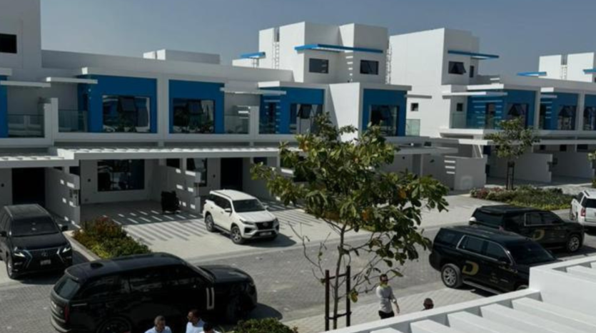 Un quartier résidentiel moderne de Dubaï avec des maisons contemporaines blanches et bleues, chacune dotée de toits plats et de balcons. La rue est bordée de voitures garées et de quelques passants. Les arbres ajoutent de la verdure à