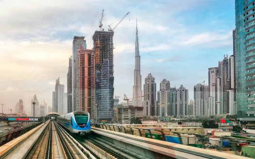 Une rame de métro de Dubaï circule sur des voies surélevées avec en toile de fond les toits de la ville, avec des gratte-ciel modernes et des grues de construction sous un ciel nuageux, soulignant les opportunités d&#039;investissement à Dubaï.