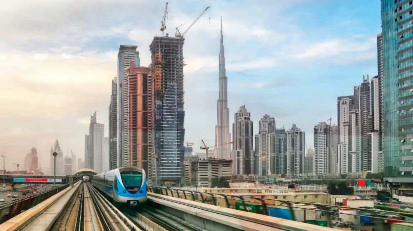 Une rame de métro de Dubaï circule sur des voies surélevées avec en toile de fond les toits de la ville, avec des gratte-ciel modernes et des grues de construction sous un ciel nuageux, soulignant les opportunités d'investissement à Dubaï.