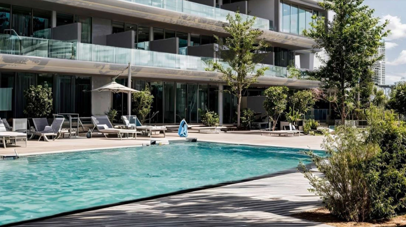 Espace piscine extérieure moderne avec chaises longues et parasols à côté d&#039;un immeuble résidentiel de plusieurs étages doté de vastes balcons et de grandes façades vitrées sous un ciel bleu clair à Dubaï.
