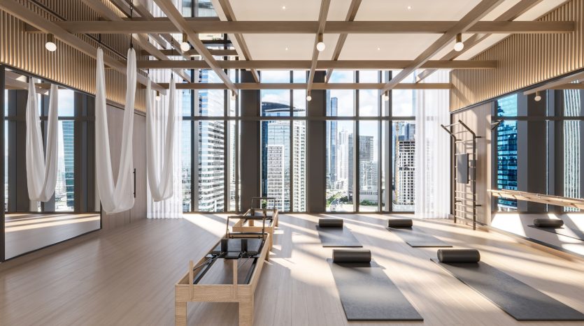 Studio de yoga moderne avec parquet et plafond en bois, grandes fenêtres avec vue sur la ville de Dubaï, tapis au sol et mobilier minimaliste sous une lumière naturelle vive.