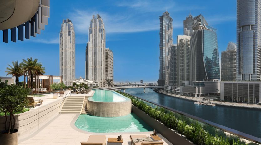 Une vue panoramique sur une luxueuse terrasse de piscine avec chaises longues, surplombant un paysage urbain moderne avec de grands gratte-ciel et une rivière à Dubaï, dans un cadre lumineux et ensoleillé.