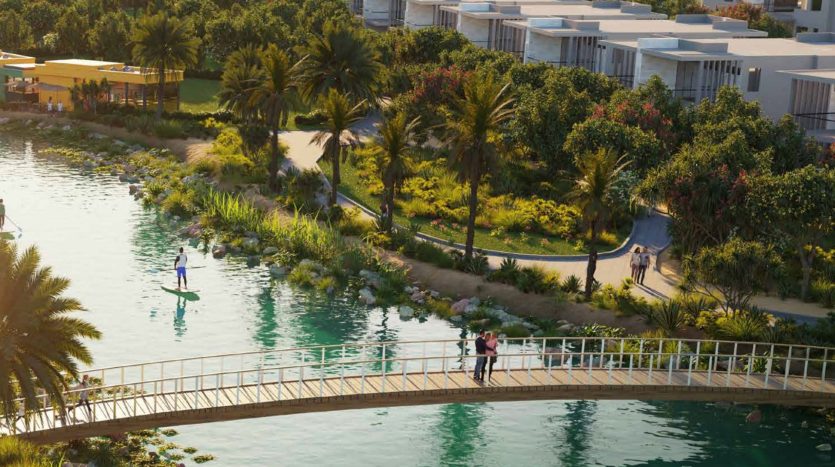 Une vue panoramique sur un parc luxuriant avec des gens marchant et faisant du paddleboard le long d&#039;une rivière, avec un pont incurvé et des immeubles d&#039;appartements modernes à Dubaï en arrière-plan.