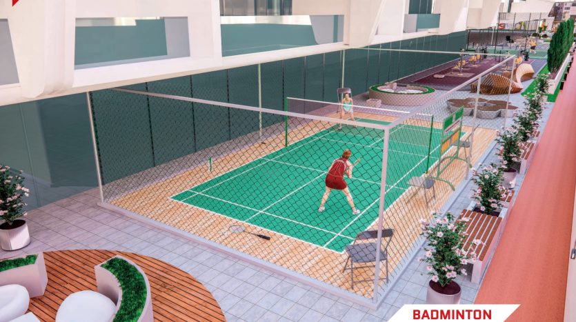 Une personne en tenue rouge jouant au badminton sur un terrain vert dans un appartement sur le toit de Dubaï, entouré de barrières de verre, de plantes et de mobilier d&#039;extérieur, avec des bâtiments en arrière-plan.