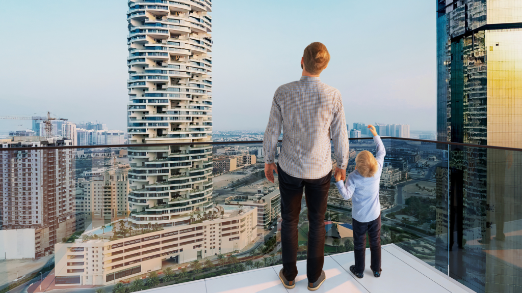 Un homme et un jeune enfant, tous deux regardant un paysage urbain depuis le haut balcon d'un appartement à Dubaï, avec des gratte-ciel modernes et des chantiers de construction visibles en arrière-plan.