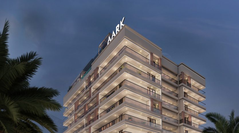 Bâtiment moderne de plusieurs étages au crépuscule avec panneau « arche » sur le toit, entouré de palmiers à Dubaï. Le bâtiment dispose de lumières intérieures et de balcons lumineux.