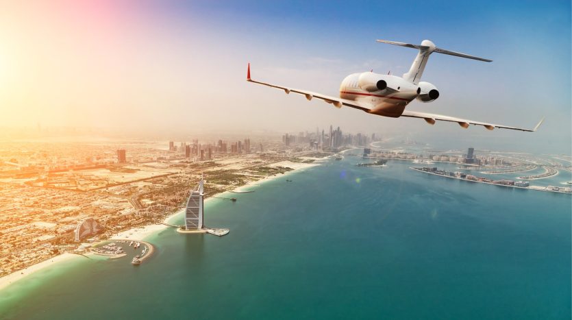 Un jet privé survolant une ville côtière ensoleillée avec des gratte-ciel, des monuments emblématiques et le premier immobilier Dubaï visible, mettant en valeur un ciel clair et des eaux chatoyantes.
