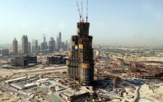 Vue grand angle d'un grand chantier de construction à Dubaï avec la structure squelettique d'un immeuble imposant au premier plan, entouré de divers autres immeubles de grande hauteur en construction, sous un ciel brumeux.