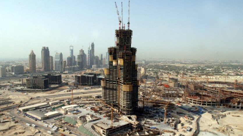 Vue grand angle d&#039;un grand chantier de construction à Dubaï avec la structure squelettique d&#039;un immeuble imposant au premier plan, entouré de divers autres immeubles de grande hauteur en construction, sous un ciel brumeux.
