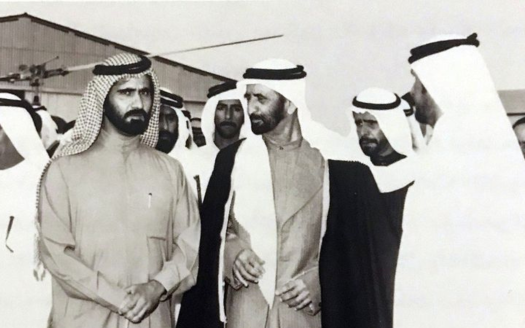 Photo en noir et blanc d'un groupe d'hommes du Moyen-Orient en tenue traditionnelle engagés dans une conversation sur l'immobilier Dubaï, debout à l'extérieur avec des bâtiments visibles en arrière-plan.