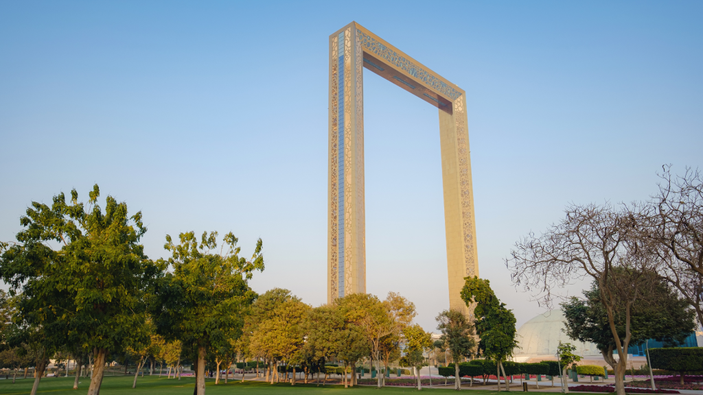 Une grande structure rectangulaire en forme de cadre doré se dressant dans un parc avec des arbres verts et des pelouses bien entretenues sous un ciel bleu clair, symbolisant les opportunités d&#039;investissement luxueuses à Dubaï.