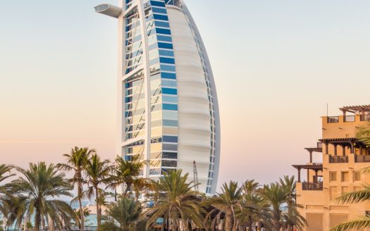 Vue de l&#039;hôtel Burj Al Arab à Dubaï au coucher du soleil, encadrée par des palmiers et l&#039;architecture de la villa adjacente de Dubaï, sous un ciel rose tendre.