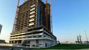 Un grande edificio in costruzione a Dubai, con diversi piani e impalcature, su uno sfondo di cielo sereno. In primo piano un'area erbosa e altri edifici in lontananza.