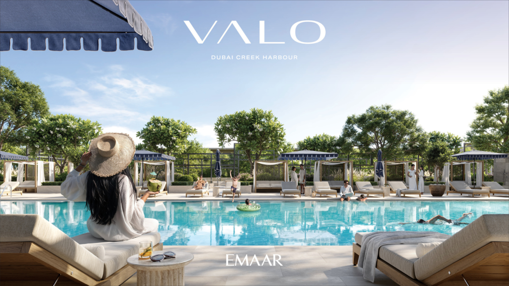 Une femme portant un chapeau de soleil blanc sirote un verre tout en surplombant une piscine luxueuse entourée de palmiers et de chaises longues sous un ciel clair, avec le texte « VALO Dubai Creek Harbour by EMAAR Immobilier Dubai » en haut.