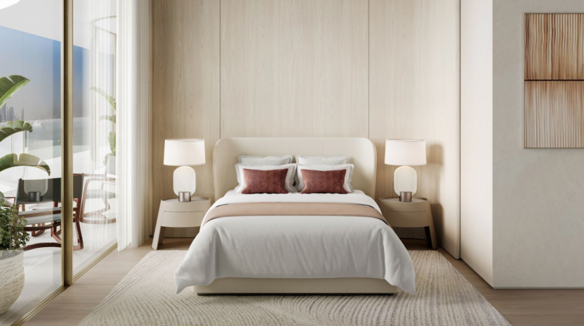 Une chambre moderne du Dubai Harbour Residences comprend un grand lit centré contre un mur en bois clair. Le lit, fait de draps blancs et beiges, dispose de deux oreillers décoratifs marron. Des tables de chevet assorties avec des lampes sont placées des deux côtés, tandis qu'une plante en pot et de vastes fenêtres ornent la gauche.