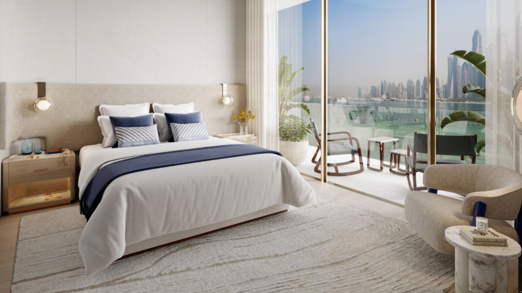 Une chambre d'hôtel moderne au Dubai Harbour Residences dispose d'un grand lit avec des draps blancs et des touches bleues. La chambre comprend une table de nuit, un fauteuil et un balcon avec coin salon extérieur donnant sur les toits de la ville et le paysage aquatique. Les plantes en pot ajoutent une touche de verdure à l’atmosphère relaxante.