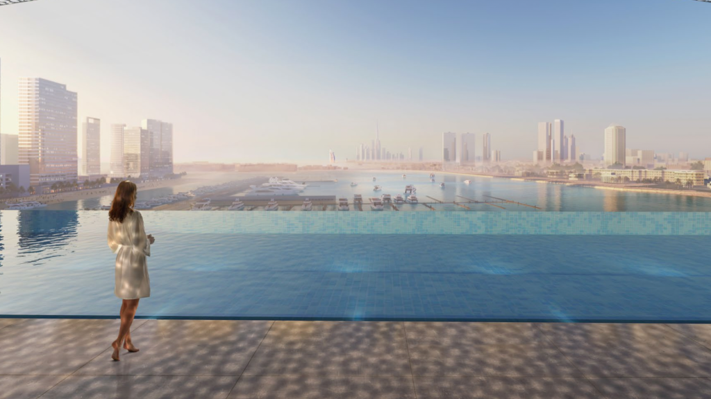 Une femme vêtue d'une robe blanche se tient au bord d'une piscine à débordement et regarde l'horizon moderne des Dubai Harbour Residences avec ses grands immeubles et ses aménagements en bord de mer pendant la journée. L'eau de la piscine se fond harmonieusement avec l'horizon lointain.