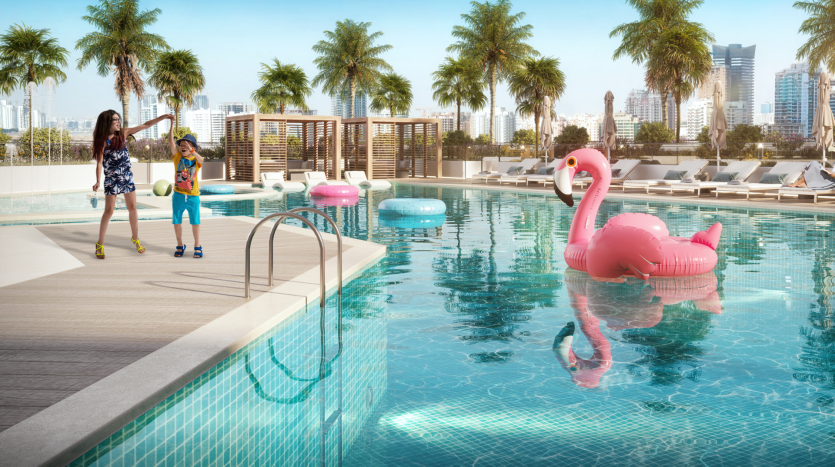 Une mère et son enfant interagissent de manière ludique au bord d&#039;une piscine ensoleillée sur le toit avec un grand flamant rose flottant à Dubaï. Paysage urbain moderne en arrière-plan avec un ciel clair.