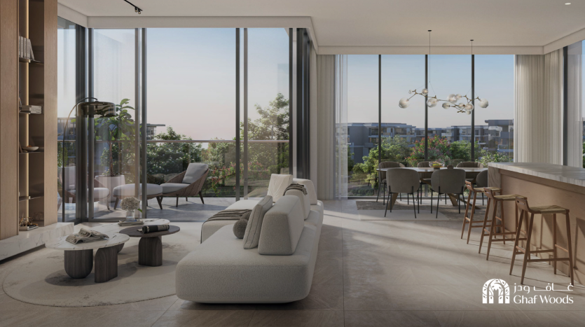 Un salon et salle à manger moderne à aire ouverte comprend un élégant canapé blanc, une table basse minimaliste, des baies vitrées, un lustre contemporain et un comptoir de bar avec des tabourets. À l’extérieur, le grand balcon du Ghaf Woods surplombe un paysage verdoyant.