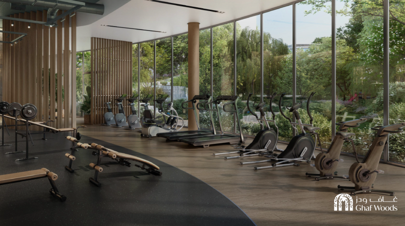 Une salle de sport moderne avec des baies vitrées donnant sur un jardin luxuriant à l'extérieur. La salle de sport est équipée de diverses machines d'entraînement, notamment de tapis roulants, de vélos d'exercice et d'équipements d'haltérophilie. Le logo « Ghaf Woods » est visible dans le coin inférieur droit, se fondant parfaitement dans l'environnement serein.