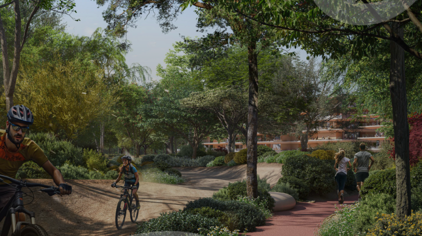 Un parc pittoresque avec un chemin de gravier et une piste de course rouge entouré de bois de ghaf luxuriants. Deux cyclistes roulent sur le chemin de gravier, tandis que deux personnes courent sur la piste de course. Les arbres et les buissons fournissent suffisamment d'ombre et un bâtiment est visible à l'arrière-plan.