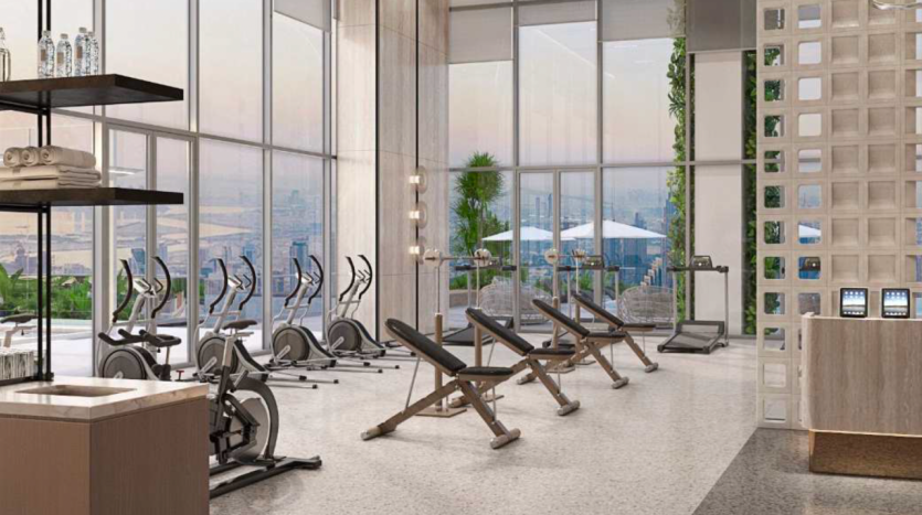 Salle de sport moderne avec appareils de cardio-training et bancs de musculation, baies vitrées donnant sur un paysage urbain et station d'eau sur la gauche dans un appartement de Dubaï.