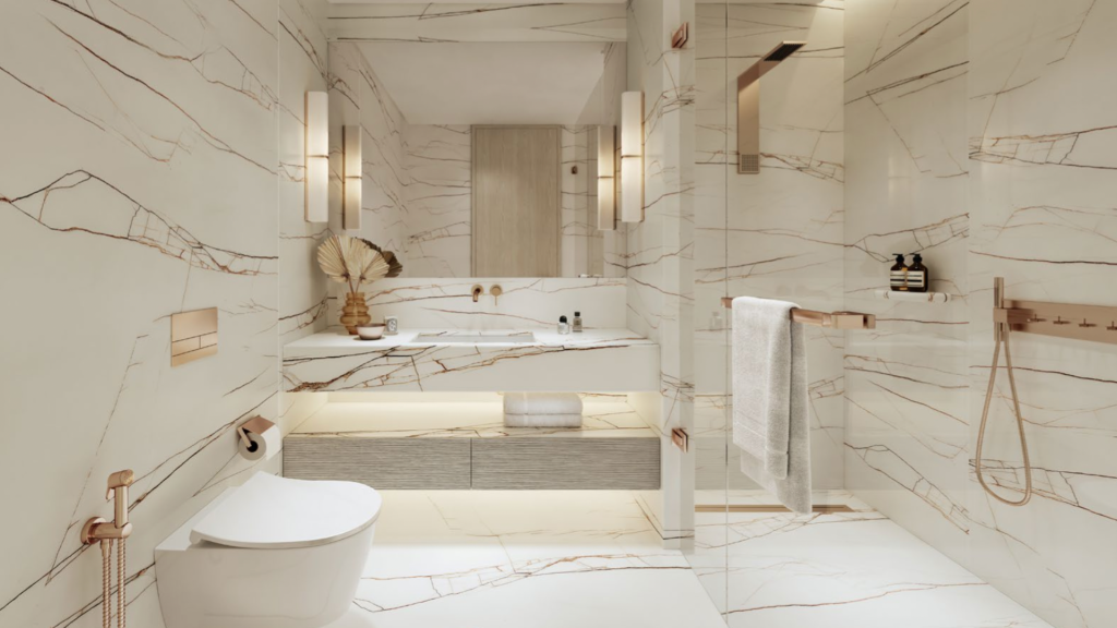 Salle de bains moderne et luxueuse des Dubai Harbour Residences avec murs et sols en marbre beige, comprenant des toilettes blanches suspendues, un grand miroir, deux lavabos, une douche à l'italienne avec porte vitrée et d'élégants luminaires métalliques. Une serviette est suspendue à un rail et des objets de décoration sont placés près de l'évier.