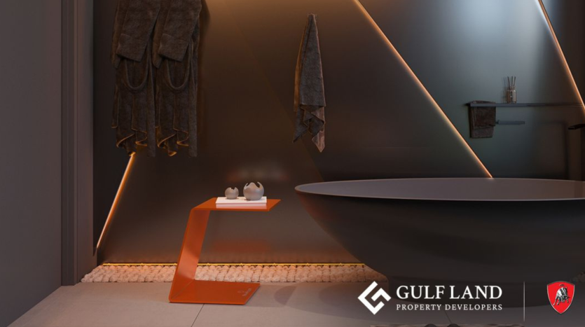 Une salle de bain moderne avec des murs sombres et un éclairage élégant. Une baignoire ronde repose sur une base de galets blancs à côté d’une table d’appoint angulaire orange et de trois serviettes sombres accrochées au mur. Le logo « Gulf Land Property Developers », associé à Tonino Lamborghini Residences Dubai, est visible en bas à droite.