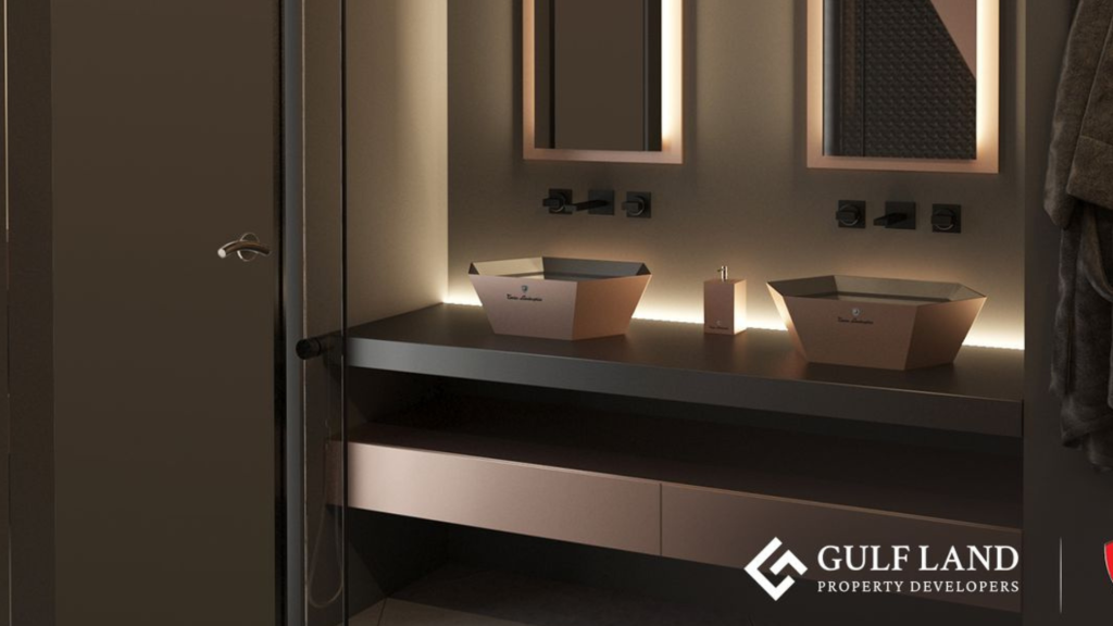 Une salle de bains moderne avec un éclairage ambiant doux comprend un comptoir noir élégant avec deux lavabos de forme carrée. Il y a deux miroirs au-dessus des lavabos et des crochets à serviettes sur le mur de droite. Le logo « Gulf Land Property Developers » se trouve dans le coin inférieur droit, faisant allusion à Tonino Lamborghini Residences Dubai.