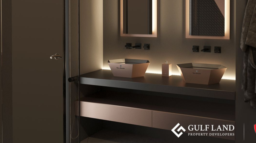 Une salle de bains moderne avec un éclairage ambiant doux comprend un comptoir noir élégant avec deux lavabos de forme carrée. Il y a deux miroirs au-dessus des lavabos et des crochets à serviettes sur le mur de droite. Le logo « Gulf Land Property Developers » se trouve dans le coin inférieur droit, faisant allusion à Tonino Lamborghini Residences Dubai.