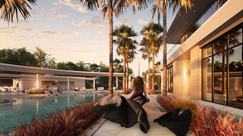 Une femme se détend sur une chaise longue surplombant une piscine luxueuse entourée de palmiers au coucher du soleil à la Villa Karl Lagerfeld. Sa vue inclut l'eau sereine et la façade moderne du bâtiment.