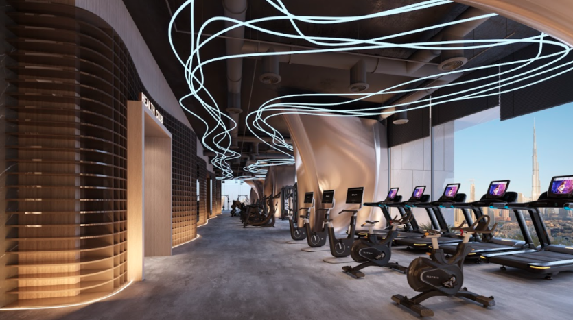 Une salle de sport moderne comprend des tapis roulants, des vélos d'exercice et d'autres équipements de fitness disposés dans un intérieur élégant et spacieux. L'éclairage incurvé accentue le plafond et les grandes fenêtres offrent une vue sur un paysage urbain avec un gratte-ciel proéminent au loin.