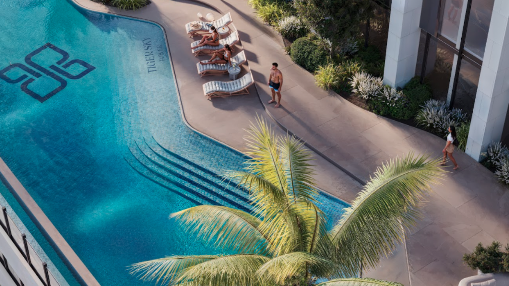 Vue aérienne d'une piscine avec une terrasse incurvée et plusieurs chaises longues, certaines occupées par des personnes prenant un bain de soleil. Un homme en maillot de bain et une femme avec un chapeau marchent sur la terrasse de la piscine. Un palmier projette une ombre près de la piscine et de la verdure entoure la zone.