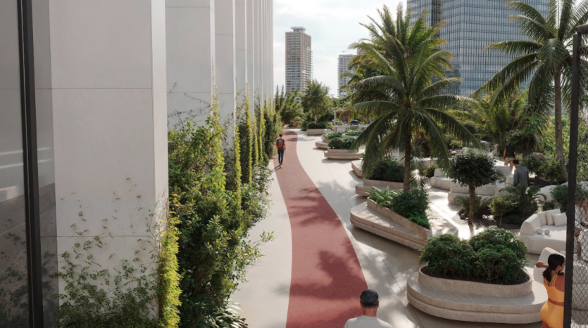 Une allée urbaine moderne bordée de verdure, comprenant de grands palmiers et arbustes, longe un bâtiment contemporain. Le chemin comporte une piste de marche rouge et est peuplé de quelques piétons. En arrière-plan, des immeubles de grande hauteur sont visibles.