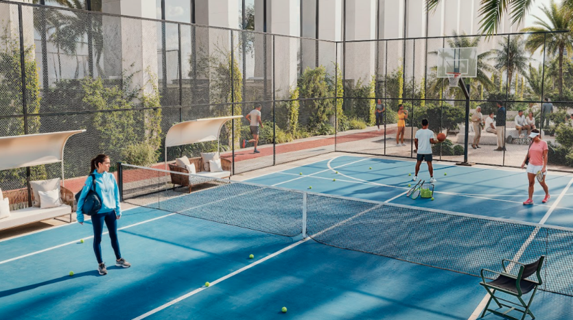 Des personnes participant à diverses activités sur un terrain bleu polyvalent entouré de hauts filets. Certains jouent au tennis, tandis que d’autres tirent au basket. La cour est flanquée de bancs blancs et de sièges rembourrés. Une verdure luxuriante et de hautes fenêtres en verre forment la toile de fond.