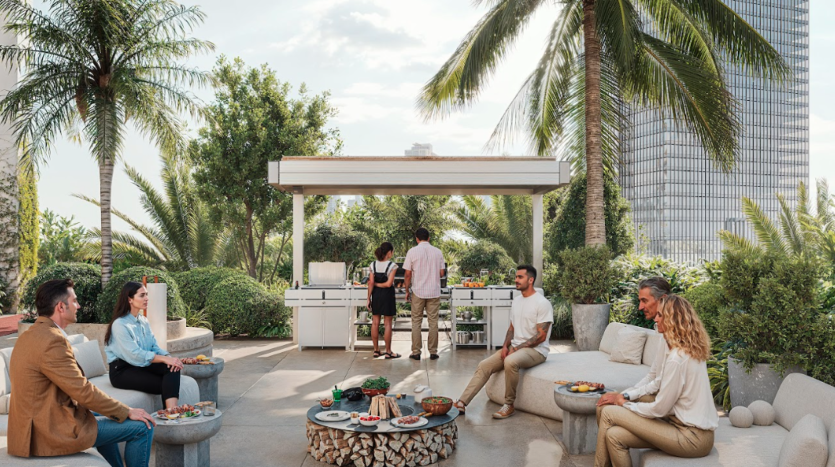 Un groupe de six personnes profite d’un rassemblement en plein air dans un jardin luxuriant planté de palmiers. Ils sont assis sur des bancs et des tabourets autour de tables avec de la nourriture, tandis que deux individus cuisinent dans une cuisine extérieure en arrière-plan. Un grand bâtiment est visible derrière.