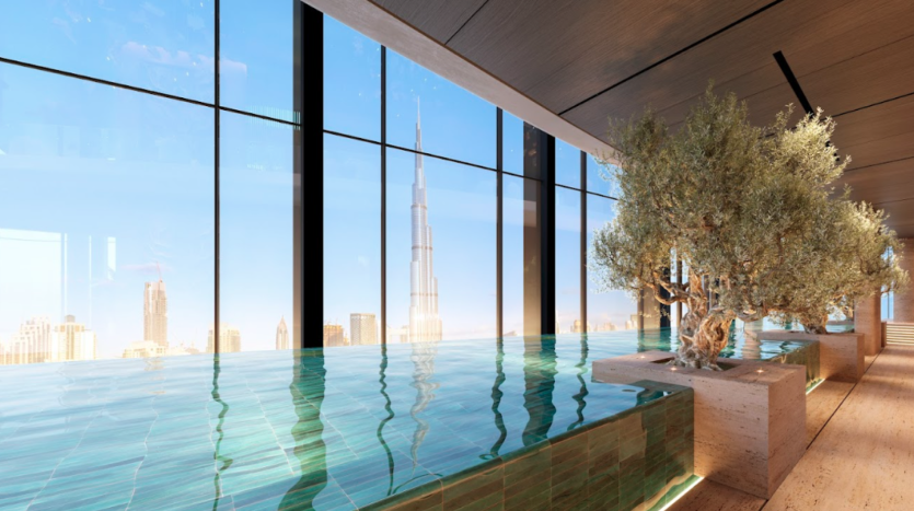 Une piscine intérieure à débordement avec vue sur les toits de la ville grâce à de grandes baies vitrées. Un grand gratte-ciel se détache en arrière-plan. Des oliviers dans des jardinières beiges bordent la terrasse en bois à côté de la piscine. L'espace est bien éclairé par la lumière naturelle.