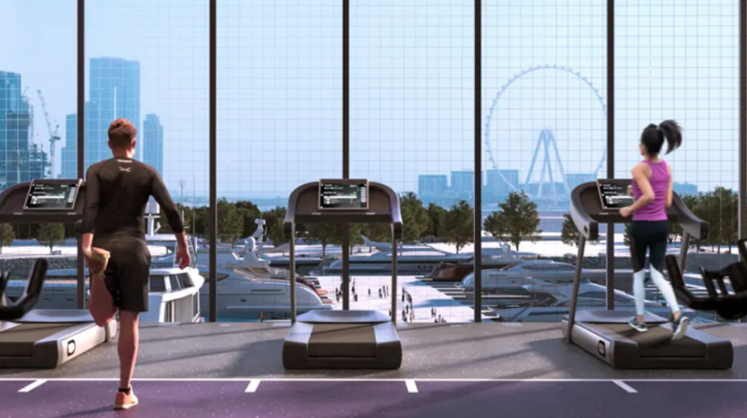 Deux personnes courent sur des tapis roulants dans une salle de sport moderne dotée de grandes fenêtres. La vue extérieure révèle un paysage urbain avec des gratte-ciel, de la verdure, des bateaux et une grande roue en arrière-plan. L’homme à gauche porte du noir et la femme à droite porte du rose.