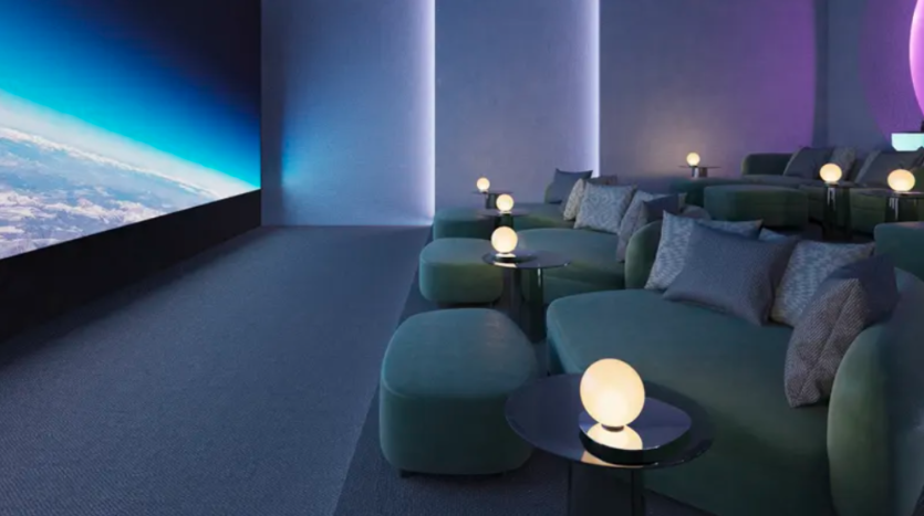 Un cinéma maison moderne et luxueux avec des fauteuils inclinables et des poufs verts moelleux. Chaque coin salon dispose d'une petite table avec une lampe ronde. Un grand écran affichant une image de l'espace avec la Terre visible domine le mur opposé au coin salon.