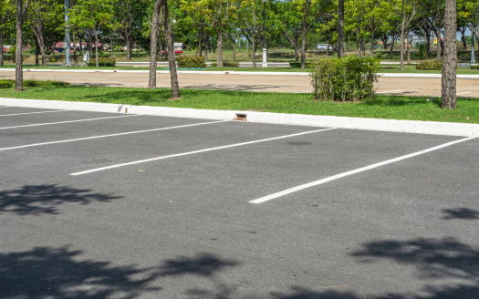 Un parking vide avec des lignes blanches fraîchement peintes, également appelés places de parking, est entouré d'herbe verte, d'arbres et de quelques buissons. Les ombres des arbres tombent sur l'asphalte. Quelques voitures et une route sont visibles en arrière-plan au-delà de l'herbe. Le décor évoque un coin étonnamment serein de Dubaï.