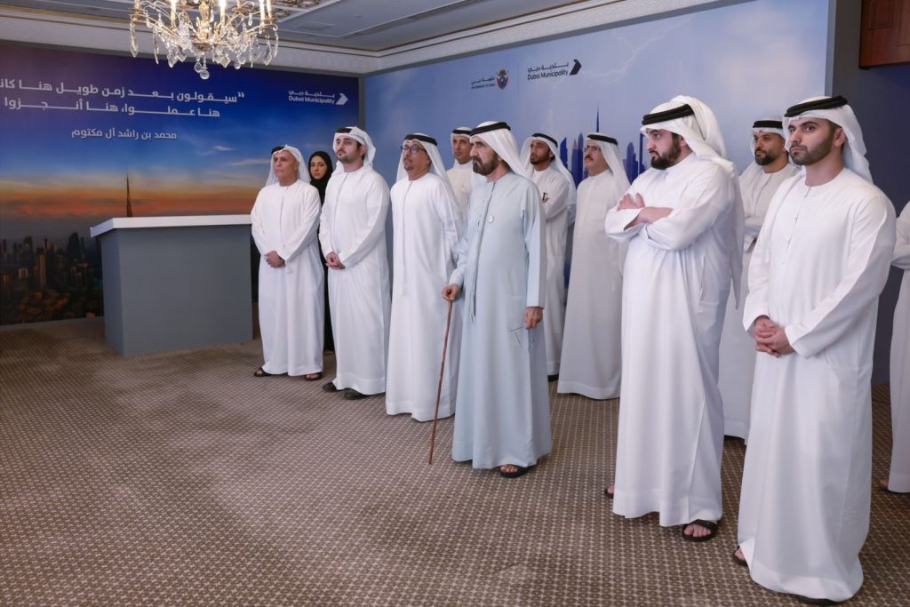 Un groupe d’hommes en tenue traditionnelle du Moyen-Orient se tiennent dans une pièce au fond bleu et orange. Un homme parle à l’aide d’une canne tandis que d’autres écoutent attentivement. Un lustre est suspendu au plafond et une femme se tient parmi le groupe à l'arrière-plan, discutant du système d'égouts de Dubaï.