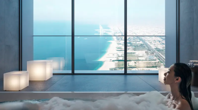 Une personne se détend dans une baignoire moderne remplie de bulles, face à une grande baie vitrée offrant une vue imprenable sur le littoral, la ville tentaculaire à côté et la mer bleue s'étendant jusqu'à l'horizon sous un ciel clair.